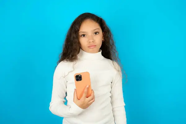 青い背景の上に白いタートルネックを身に着けている不満の美しい子供の少女はモバイルソフトウェアのアプリケーションとインターネット上のシェフの情報を使用し 現代のモバイルハンドを保持します — ストック写真