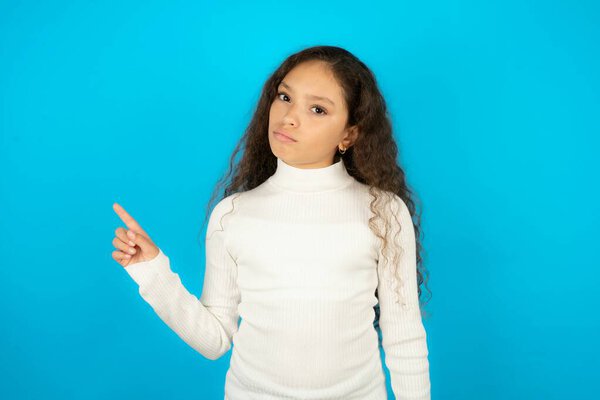 Serious Beautiful kid girl wearing white turtleneck over blue background smirks face points away on copy space shows something unpleasant. Look at this advertisement. Big price concept