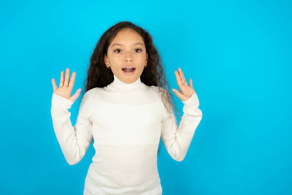 Delighted positive Beautiful kid girl wearing white turtleneck over blue background opens mouth  and arms palms up after having great result