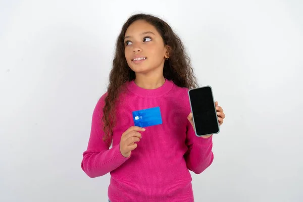 白い背景の上にピンクのタートルネックを身に着けている美しい少女 銀行カードを保持する現代装置 空のスペースを見る — ストック写真