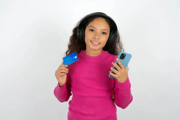 漂亮的小女孩穿着粉色的高领毛衣 白色背景 耳朵上戴着立体声耳机 手持现代手机和信用卡 — 图库照片