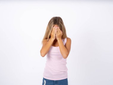 Beyaz stüdyo arka planında kolsuz tişört giyen sarışın kız yüzünü elleriyle kapatıyor, yıkılmış ve ağlıyor. Üzücü bir kavram