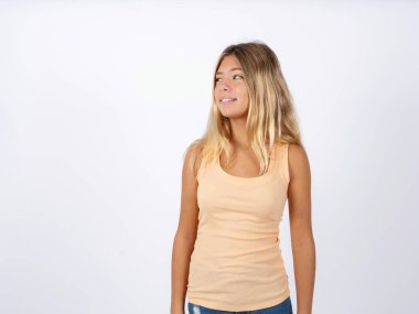 Beyaz stüdyo arka planında kolsuz tişört giyen, dudak ısıran ve boşluğu boşaltmak için kurnaz görünen sarışın genç kız.. 