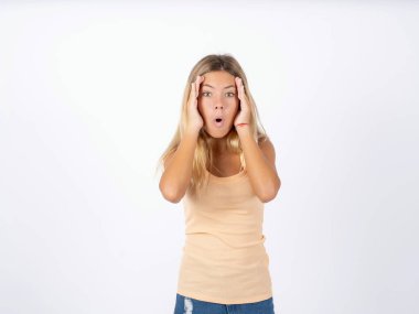 blonde teen girl wearing sleeveless t-shirt over white studio background with scared expression, keeps hands on head, jaw dropped, has terrific expression. Omg concept clipart