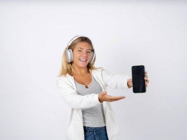 Kulaklık takan, beyaz ceketli, cep telefonlu genç bir kız. Akıllı telefonu sunuyorum. Reklam kavramı. 