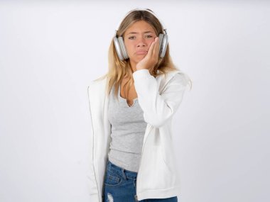 Sad lonely teen girl with headphones wearing white jacket over white studio background touches cheek with hand bites lower lip and gazes with displeasure. Bad emotions clipart