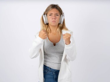 Displeased annoyed teen girl with headphones wearing white jacket over white studio background clenches fists, gestures pissed, ready to revenge, looks with aggression at camera stands full of hate, being pressured clipart