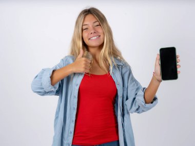 Beyaz stüdyo arka planında kot ceket giyen sarışın genç kız boş ekran akıllı telefonu göster, başparmağını kaldır ve yeni bir uygulama tavsiye et 