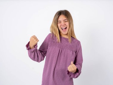 beautiful teenager girl wearing purple T-shirt over white studio background very happy and excited doing winner gesture with arms raised, smiling and screaming for success. Celebration concept. clipart