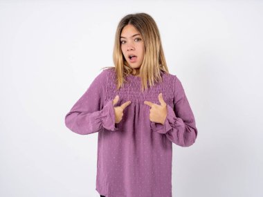 Embarrassed beautiful teenager girl wearing purple T-shirt over white studio background indicates at herself with puzzled expression, being shocked to be chosen to participate in competition, hesitates about something  clipart