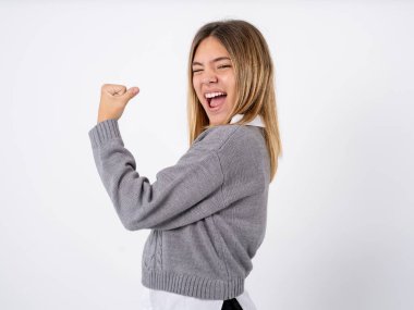 Profil yan görüntüsü beyaz stüdyo geçmişi üzerine gri süveter giyen genç kız zaferi kutluyor