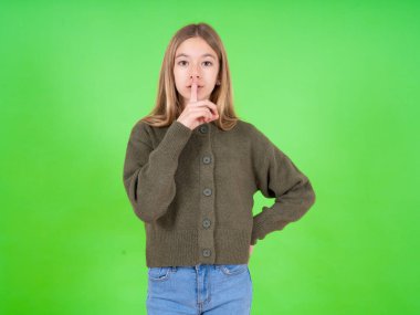 Yeşil kazak ve kot pantolon giyen güzel beyaz kız sessiz jest yapıyor, parmaklarını dudaklarından üstün tutuyor. Sessizlik ve gizli konsept.