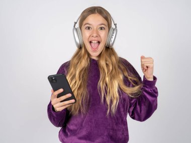 Mor kazak giyen olumlu genç kız, kulaklığa bağlı modern cep telefonunu tutuyor, iyi duygulardan yumruğunu sıkıyor, sevinçle haykırıyor., 