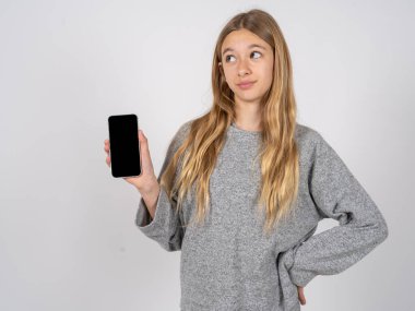 Gri eşofman giyen güzel genç kız yeni bir cep telefonu tutuyor ve gizemli görünüyor. Modern cep telefonunun boş görüntülerini gösteriyor.