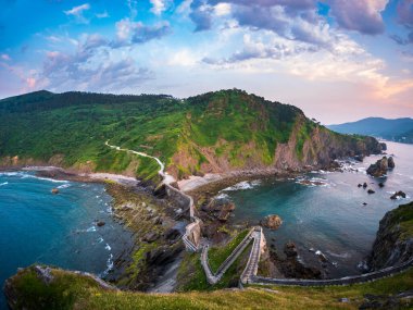Gaztelugatxe 'nin arkasında gün batımı Bask Ülkesi İspanya dolambaçlı taş köprüsü inzivaya çekilen kayalık adaya doğru tırmanıyor sakin deniz parlayan gökyüzü antik yol mistisizm inanç sonsuz yalnızlık yolculuğu