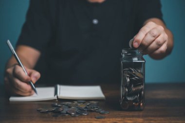 Genç adam kavanoza para koyar ve not defterine rekor miktarda para koyar, biriktirme, hayır kurumu, aile finans planı konsepti, bağış toplama, üst yönetim, yatırım, finansal kriz konsepti