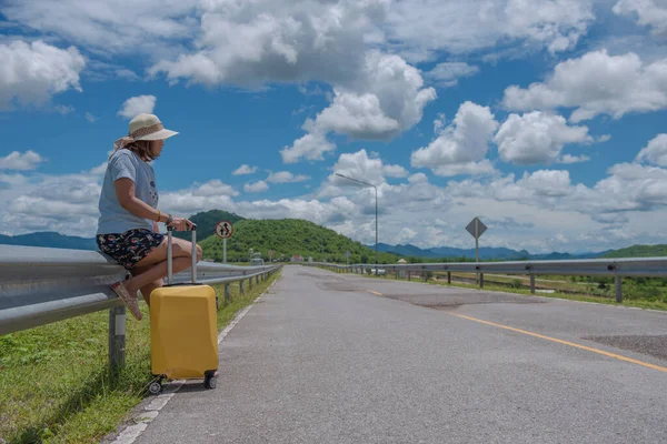 青い空と美しい自然の風景の中で車を待っている旅行スーツケースと道路上のヒッチハイクアジアの女性 晴れた日の旅行のコンセプト — ストック写真