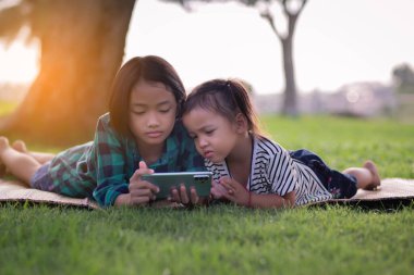 Çimenlerde uzanmış telefonlarına bakan iki genç kız, yaz, altın saat, gün batımı. SSTKHome