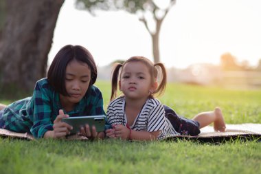 Çimenlerde uzanmış telefonlarına bakan iki genç kız, yaz, altın saat, gün batımı. SSTKHome