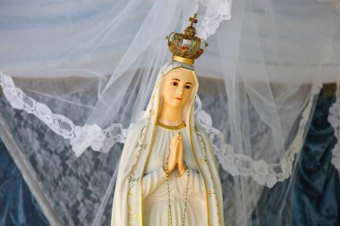 Taçla süslenmiş Bakire Meryem heykeli ve narin beyaz bir dantel peçenin altında dua ediyor.