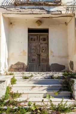 Terk edilmiş bir evin ön cephesinde eski ahşap bir kapı. Merdivenlerde bitkiler yetişiyor.