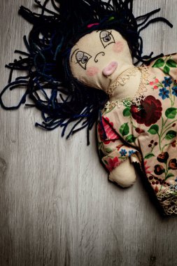 Mavi iplik saçlı yaşlı bez bebek kırsal bir ahşap zeminde huzur içinde yatıyor, çocukluk anılarını ve nostaljiyi anımsatıyor. Çiçekli elbiseli bebek yalnızlık yayar, zamanın akışını sembolize eder.