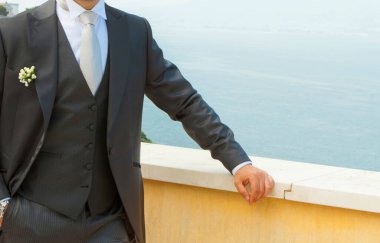 Damat, yaka çiçekli bir takım elbise giyiyor. Arka planda okyanus olan korkuluklara yaslanıyor.