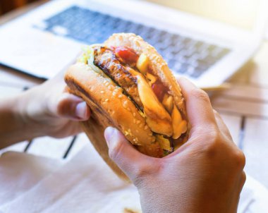 Elleri lezzetli bir burger, patates kızartması ve erimiş peynirle arka planda bulanık bir dizüstü bilgisayar tutuyor.