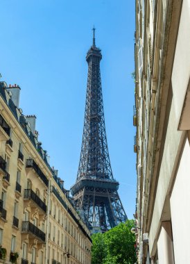 Eyfel Kulesi, güneşli bir günde klasik apartman binalarıyla kaplı dar bir Paris caddesinin üzerinde yükseliyor.