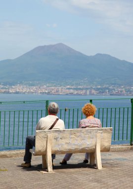 Emekli çift güneşli bir günde bir banktan Vezüv Yanardağı ve Napoli Körfezi manzarasının keyfini çıkarıyorlar.