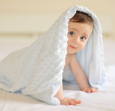 Sevimli erkek bebek, yumuşak mavi bir battaniyenin altında, bir yatakta emekliyor, rahatlığın ve oyunculuğun tadını çıkarıyor.