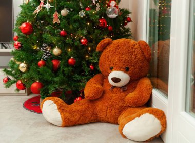 Süslü bir Noel ağacının yanında oturan büyük kahverengi ayıcık rahat ve şen bir atmosfer yaratıyor.