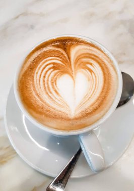 Lezzetli bir fincan kapuçinonun tadını çıkarıyorum. Güzel kalp şeklinde latte sanatı, beyaz bir fincan ve mermer bir masada servis edilen çay tabağı.