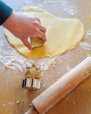 Çocuk hamurun üzerinde kalp şeklinde kurabiye keskisi, merdane ve ahşap masa üzerinde yıldız şeklinde kesiciler kullanarak kurabiye yapıyor.