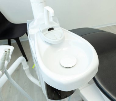 Diş kliniğindeki bir dişçi koltuğunun musluğu ve bardak tutacağı ile durulamaya hazır beyaz lavabo.