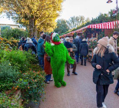 Castelfalfi, İtalya - 15 Aralık 2024: Grinch kostümlü bir kişi Noel alışverişinde ziyaretçilere el sallıyor, bayram neşesini yayıyor.