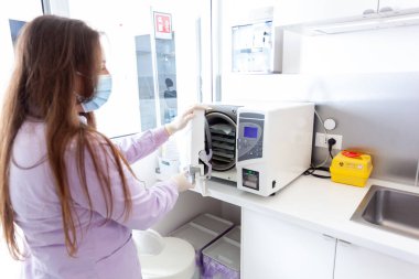 Maske ve eldiven takan hemşire modern bir klinikte tıbbi aletleri sterilize etmek için otoklav kullanıyor.