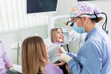 Aynaya bakan kadın dişçi, dişçi kliniğindeki tatmin olmuş hastanın tedavisinin sonucunu gösteriyor.