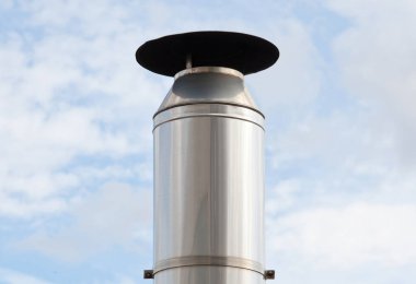 Yağmurluk takan parlak metal baca borusu bulutlu mavi gökyüzüne kirletici dumanlar salıyor.