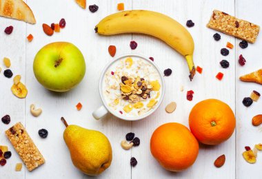 Elma, muz, armut ve portakal gibi taze meyvelerle çevrili süt ve mısır gevrekleri, kuru meyve, fındık ve enerji barları, beyaz ahşap bir masada sağlıklı bir kahvaltı hazırlıyorlar.