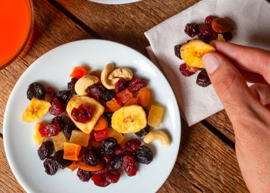 Renkli fındık ve kurutulmuş meyve karışımından elle kurutulmuş muz cipsi toplamak, ahşap bir masanın üzerinde güzel bir şekilde düzenlenmiş, yanında bir bardak canlandırıcı meyve suyu ile birlikte.