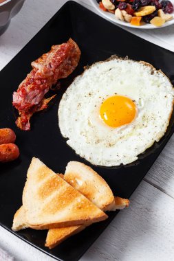 Geleneksel İngiliz kahvaltısı kızarmış yumurta, kıtır pastırma, sosis ve tostu kapsar. Siyah tabakta servis edilir ve görsel olarak çekici ve tatmin edici bir yemek oluşturur.