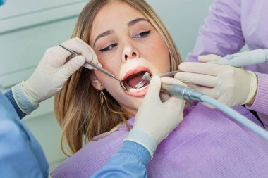 Diş hekimi, dişçi kliniğindeki genç bir kadına diş matkabı ve ayna kullanarak diş muayenesi yapıyor.