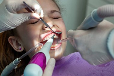 Pediatrik dişçi diş fırçası, tükürük çıkarıcı ve diş aynası kullanarak diş temizliği yapan genç bir kız üzerinde diş temizliği yapıyor.