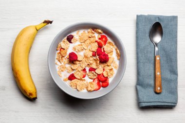 Mısır gevreği ve sütle dolu gri kase, çilek ve ahududu ile kaplı, yanında bir muz ve mavi bir peçeteyle beyaz ahşap bir masada bir kaşık