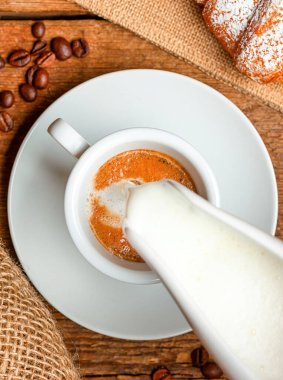 Sütlü sürahi, zengin bir kahve bardağına kremalı süt dökerek kırsal ahşap bir masada lezzetli bir macchiato yaratıyor ve yanında da gevrek bir kruvasan ve dağınık kahve çekirdekleri var.