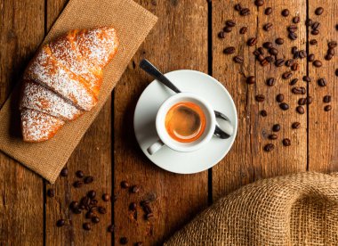 Espresso kahve fincanı gevrek bir kruvasanın yanında, kahve çekirdekleri kırsal ahşap masaya saçılmış, nefis bir İtalyan kahvaltı deneyimi için mükemmel bir ortam yaratıyor.