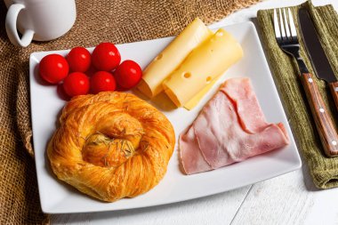 Biberiyeli brioche, peynirli jambon ve vişneli domateslerden oluşan nefis lezzetli bir kahvaltı çatal bıçaklı yeşil bir peçete ve bir fincan kahvenin yanında beyaz bir tabakta servis edilir.