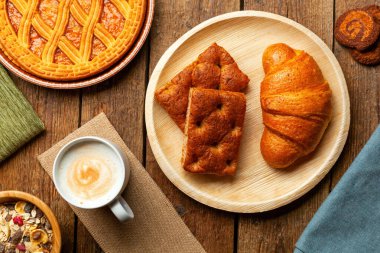 Zengin İtalyan kahvaltısı kapuçino, taze pişmiş kruvasan, schiacciata, geleneksel crostata, müsli ve ahşap bir masada kurabiyeler.