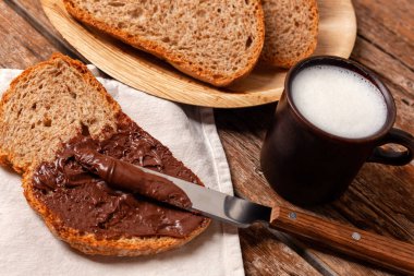 Bıçak, bir dilim kepekli ekmeğe çikolata kreması sürüyor, bir fincan süt ve arka planda daha fazla ekmek dilimleri, sağlıklı ve lezzetli bir kahvaltı hazırlıyor.